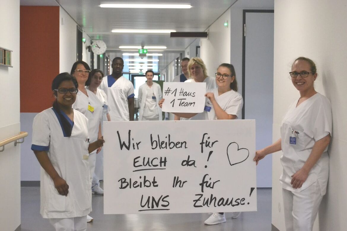 Krankenpflegerinnen und Krankenpfleger halten ein Plakat hoch, auf dem steht: Wir bleiben für euch da. Bleibt ihr für uns Zuhause!