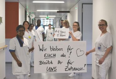 Krankenpflegerinnen und Krankenpfleger halten ein Plakat hoch, auf dem steht: Wir bleiben für euch da. Bleibt ihr für uns Zuhause!